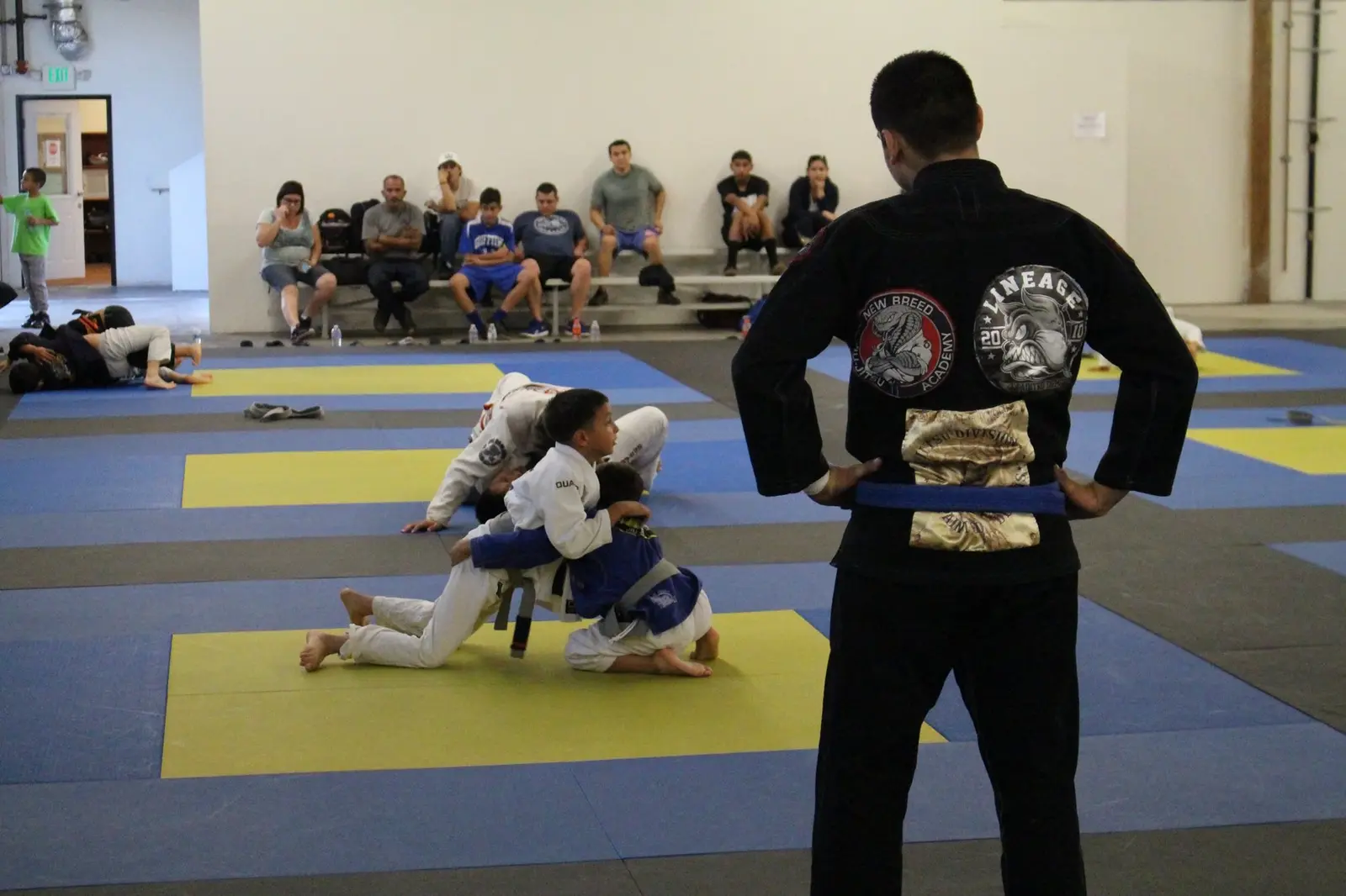 Kids practicing Jiu-Jitsu on the mat while a coach in a black gi watches